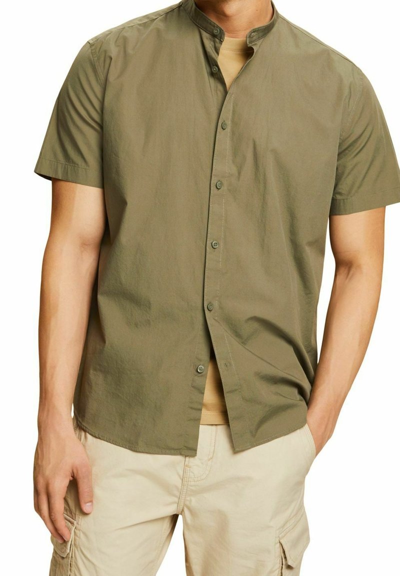 Man met olijfgroene korte mouwen overhemd met knopen over beige cargoshorts, met één hand in de zak en een neutrale achtergrond.