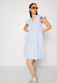 Vestido azul claro de mangas curtas com decote em V, mangas com babados e saia em camadas. Tecido suave com um corte descontraído. Usado com óculos de sol laranja.