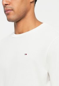 Close-up de um homem a usar uma camisola branca de gola redonda simples com um pequeno logotipo retangular em vermelho, branco e azul no peito.