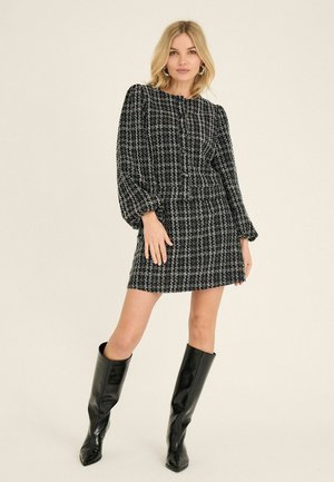 Femme aux cheveux blonds portant une mini-robe à manches longues à carreaux noirs et blancs et des bottes en cuir noires montantes jusqu'aux genoux, debout devant un fond uni.