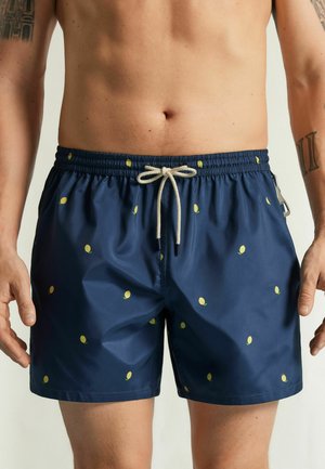 Hombre con bañador azul marino con pequeños patrones amarillos de limón y un cordón blanco en la cintura.