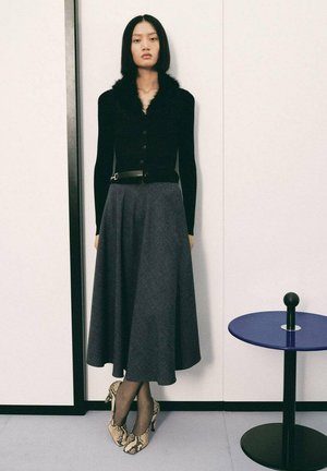 Cardigan negro con cuello de piel, falda en línea A gris, tacones de serpiente, de pie al lado de una mesa lateral circular azul. Texturas suaves, diseño moderno.
