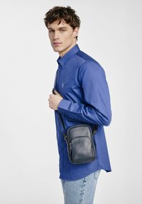 Granatowa skórzana torba crossbody o teksturowanym wykończeniu, z zamkiem błyskawicznym i regulowanym paskiem. Noszona na jasnoniebieskiej koszuli.