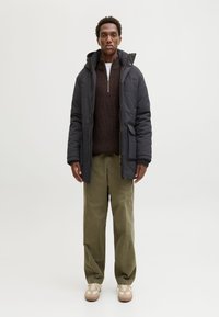 Czarna pikowana kurtka z kapturem, zapinana na zamek błyskawiczny z przodu, z bocznymi kieszeniami, noszona na brązowym swetrze dzianym, oliwkowo-zielonych spodniach i beżowych tenisówkach.