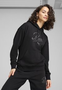Juodas megztinis su kapišonu, turintis kengūros kišenę ir sidabrinį „Puma“ logotipą. Minkšta medžiaga, laisvas fasonas ir rankogaliai.
