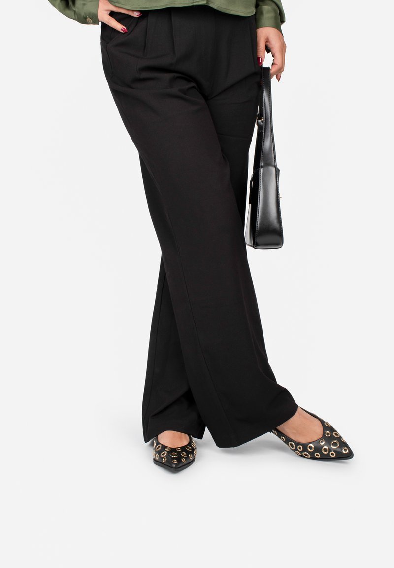 Pantaloni neri a gamba larga con pieghe e una maglietta verde; abbinati a ballerine con punta affilata con accenti in oro e una borsa nera.