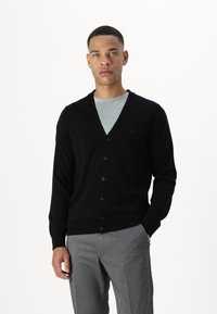 Crni kardigan dugih rukava s V izrezom i zatvaranjem na gumbe. Pleteni materijal s rebrastim manžetama i rubom. Ima mali logo na prsima.