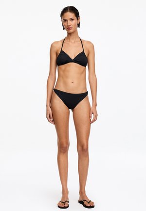 Femme debout portant un bikini halter noir et des tongs noires sur un fond blanc uni, regardant droit devant avec une expression neutre.
