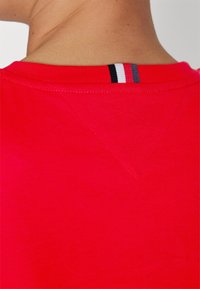 T-shirt rouge avec un col rond, doté d'une petite couture en V et d'une étiquette rayée en marine, blanc et rouge à l'arrière.