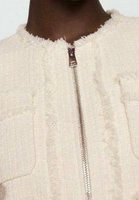 Creme tweedjakke med en central metallynlås, to brystlommer og en tekstureret finish med frynsede kanter omkring halsudskæringen.