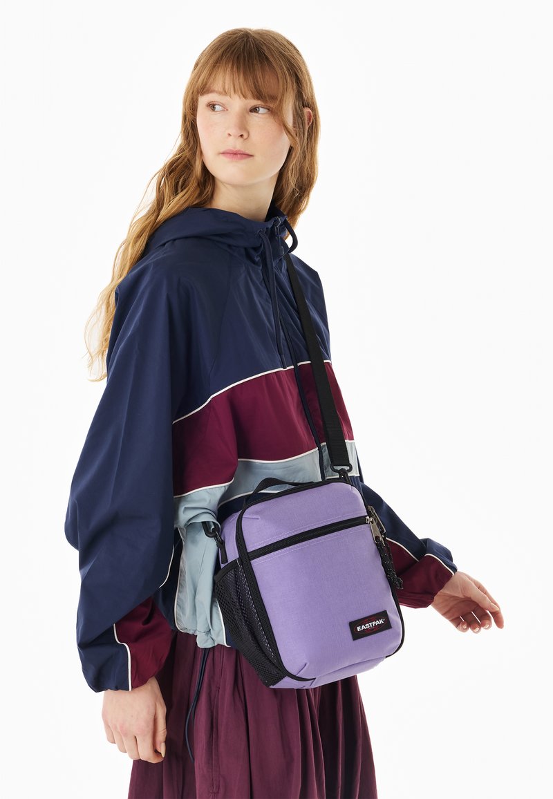 Jeune femme portant une veste à capuche bleu marine, bordeaux et bleu clair avec un sac bandoulière violet, debout devant un fond blanc.