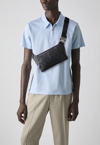 Sac à épaule noir texturé avec une finition lisse et des accessoires en métal argenté, porté en travers d'une chemise polo bleu clair.