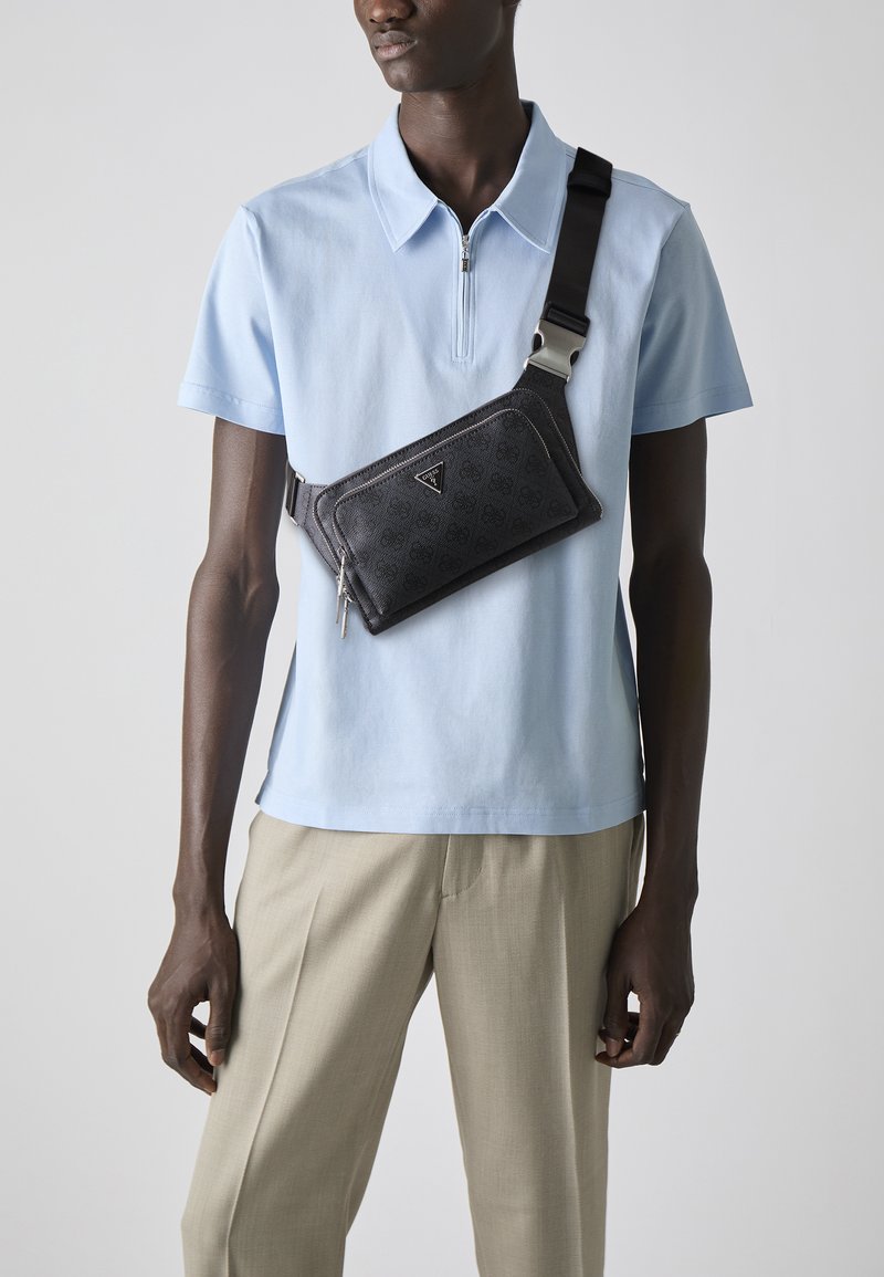 Sac à épaule noir texturé avec une finition lisse et des accessoires en métal argenté, porté en travers d'une chemise polo bleu clair.