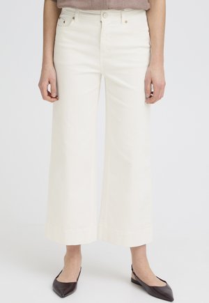 Naine, kes kannab kõrge vöökohaga, laiade säärtega valgeid pükse, mis on pahkluude kohal lõigatud, ja musti terava ninaga slingback-talle, seisab valge ühtlase tausta ees.