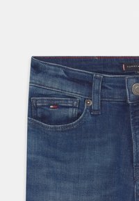 Jeans in denim di colore blu scuro con una texture liscia. Presentano una piccola tasca frontale, un bottone argentato e cuciture rosse sulla cintura.