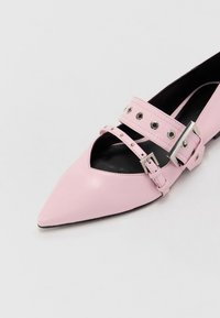 Růžová kožená botka s špičatou špičkou a černým interiérem, s dvěma pásky s hroty a velkou stříbrnou sponou na bílém pozadí.