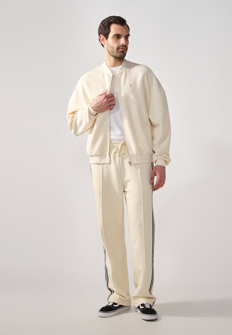 Cremefarbene Bomberjacke mit gerippter Textur, Reißverschluss und Logo auf der Brust. Passende cremefarbene Hose mit schwarzen seitlichen Streifen und Kordelzug.