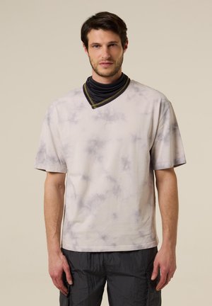 Man draagt een lichtgrijs tie-dye T-shirt met korte mouwen en een zwarte nekwarmer, staand tegen een eenvoudige beige achtergrond.
