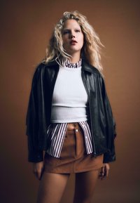 Młoda kobieta z falującymi blond włosami, ubrana w czarną skórzaną kurtkę na białym topie crop, w paskowaną koszulę i brązową zamszową mini spódniczkę, pozująca.