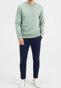 Světle zelená mikina s kulatým výstřihem a žebrovanými manžetami, se zvýrazněným textovým detailem. Spárováno s tmavě modrými slim-fit kalhotami a bílými teniskami.