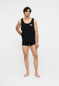 Άνδρας στέκεται ξυπόλητος με μαύρο φανελάκι με το λογότυπο "Moschino" και ταιριαστό μαύρο σορτς μπροστά σε ένα απλό λευκό φόντο.