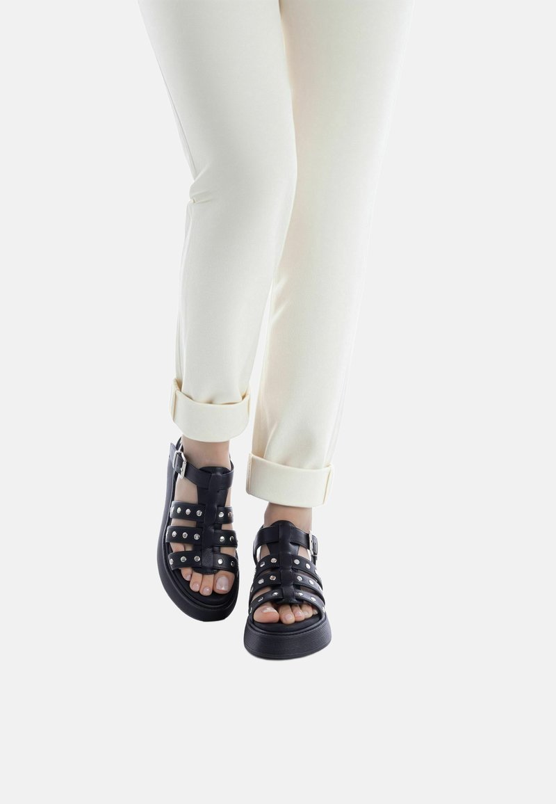 Sandalias negras con múltiples tiras y tachuelas plateadas, combinadas con pantalones claros con puños, mostrando la piel de los pies y los dedos.