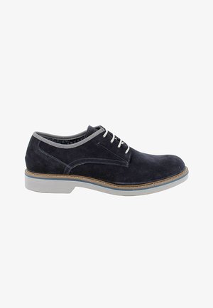 Scarpa elegante da uomo in suede nero con lacci bianchi, rifinitura grigia e suola spessa bianca con dettaglio di cuciture marroni.