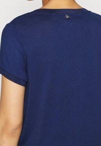 T-shirt blu navy con maniche corte, caratterizzato da un colletto testurizzato e da un piccolo logo metallico sul retro. Tessuto liscio e leggero.