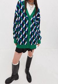 Person som bär en geometriskt mönstrad cardigan i grön, blå, svart och vit över en vit topp och svart kjol, tillsammans med svarta knähöga stövlar.