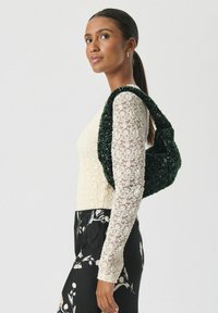 Vrouw met donker haar in een paardenstaart draagt een witte kanten top, een zwarte bloemenrok en heeft een donkergroene gestructureerde schoudertas.