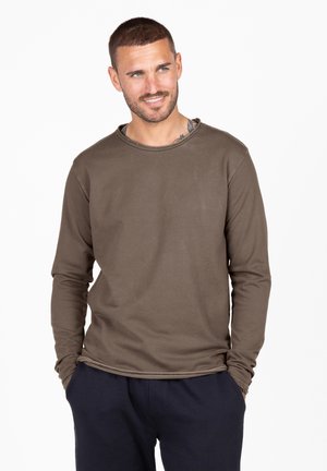 Chemise en coton marron à manches longues avec un col arrondi et des détails de couture, portée de manière décontractée sur un pantalon sombre.