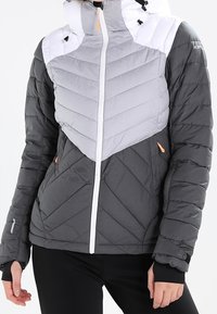 Grå och ljusgrå quiltad jacka med vit huva, med en asymmetrisk chevrondesign, orange detaljer och framzipning.