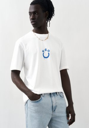 Mand med mørk hud og dreadlocks iført hvid T-shirt med blåt "HGO" smiley-design, lyseblå jeans og en perlehalskæde, poserer mod en hvid væg.