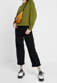 Sweat-shirt vert olive, jeans noirs amples, sac bandoulière jaune, et bottines blanches avec lacets noirs et œillets dorés.