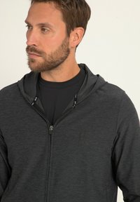 Felpa nera con zip in tessuto testurizzato, dotata di cappuccio con coulisse e un logo discreto sul petto. Indossata sopra una maglietta nera.