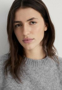 Grauer Strickpullover mit einem gerippten Ausschnitt. Besitzt eine weiche Textur, einen leichten Glanz und eine lässige Passform. Heller Hintergrund, der auf das Kleidungsstück fokussiert ist.