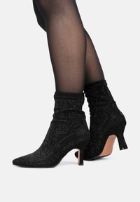 Botines de ante negro con punta afilada, diseño arrugado y adornados con tachuelas plateadas. Presentan un tacón delgado y un acabado texturizado.