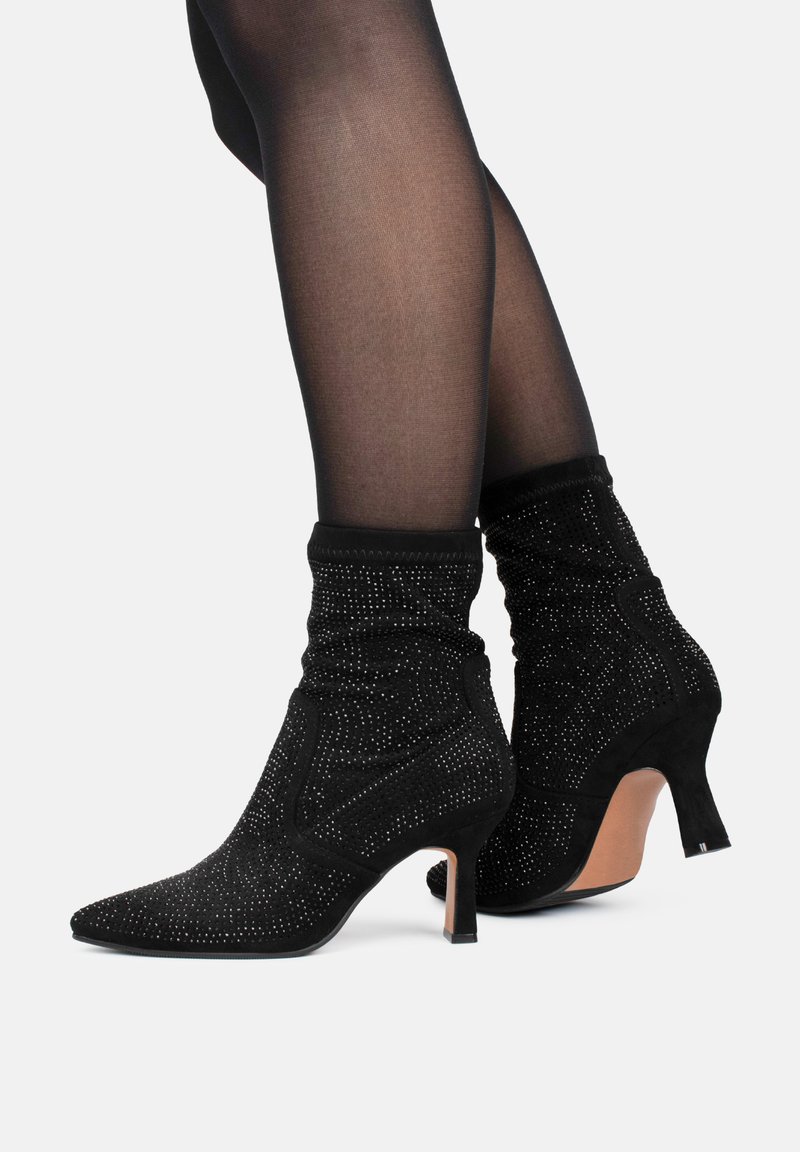 Botines de ante negro con punta afilada, diseño arrugado y adornados con tachuelas plateadas. Presentan un tacón delgado y un acabado texturizado.