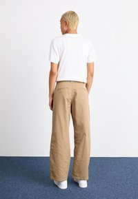 T-shirt bianca a maniche corte abbinata a pantaloni beige larghi e sneakers bianche. I pantaloni presentano due tasche posteriori e un'etichetta.