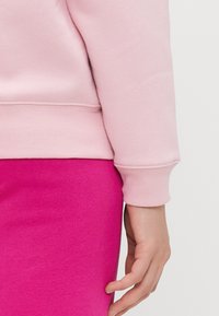 Rózsaszín pulóver bordás mandzsettákkal, élénk rózsaszín ruhadarabbal párosítva. A hangsúly a puha, texturált anyagon és a tiszta varrásokon van.