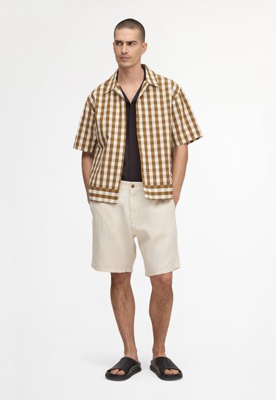 Homme portant une chemise à manches courtes marron et blanche à carreaux sur un haut noir, un short crème et des sandales noires, debout devant un fond blanc.