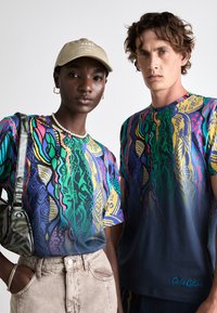 Farbenfrohe, grafische T-Shirts mit abstrakten Mustern in Blau, Grün, Gelb und Rot. Model mit beiger Mütze und Perlenkette.