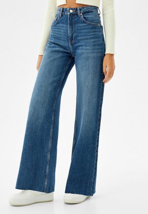 Frau trägt hochtaillierte, weit geschnittene blaue Jeans, weiße Turnschuhe und einen weißen, kurz geschnittenen Pullover mit langen Ärmeln, steht vor einem einfarbigen Hintergrund.