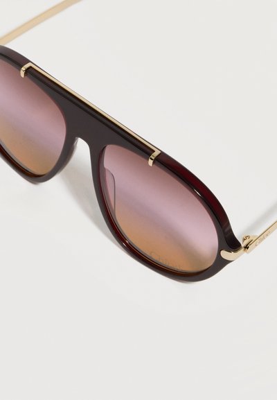 Γυαλιά ηλίου με σκούρο καφέ σκελετό, φακούς από μοβ σε κεχριμπαρένιο με διαβαθμίσεις και χρυσές λεπτομέρειες στην κορυφή και στα πλαϊνά, πάνω σε λευκό φόντο.