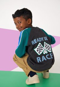 Grauer Pullover mit türkisfarbenen Ärmeln, auf der Rückseite mit dem Aufdruck "READY SET RACE" und karierten Fahnen. Helle Hose und helle Turnschuhe.