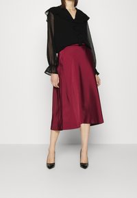 Blusa nera trasparente con colletto e polsini arricciati, abbinata a una gonna di raso bordeaux fino al ginocchio e tacchi alti neri.