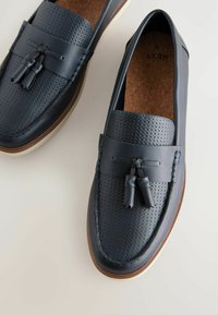 Mocasines de cuero azul con diseño perforado, acentos de borlas y suela marrón contrastante. Parte superior texturizada y acabado suave.