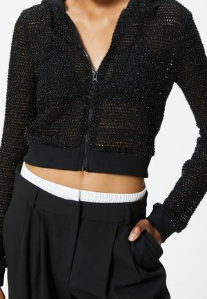 Personne portant un pull court zippé noir texturé et un pantalon noir taille haute avec une ceinture blanche visible, main dans la poche.