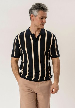 Mand med gråt hår iført en sort polo med korte ærmer med lodrette cremefarvede striber og beige bukser, der står foran en ensfarvet baggrund.