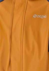 Orange vandtæt jakke med en glat tekstur, der har grå detaljer på skuldrene og et hvidt "ZIGZAG" logo på fronten.